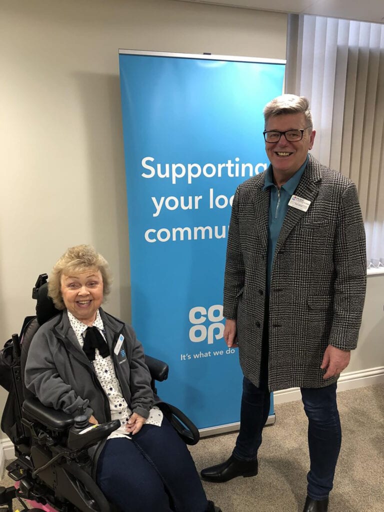 Music in Hospitals & Care volunteer Roy Jones stands next to a woman in a wheelchair in front of a co-op pull up banner.
