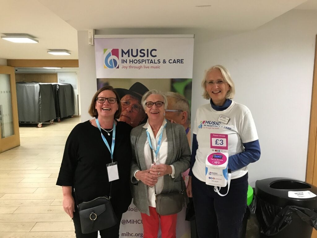 Music in Hospitals & Care Fundraiser Sue, volunteer Gillian and Chair Maureen stand in front of a pull up banner with a contactless donation machine.