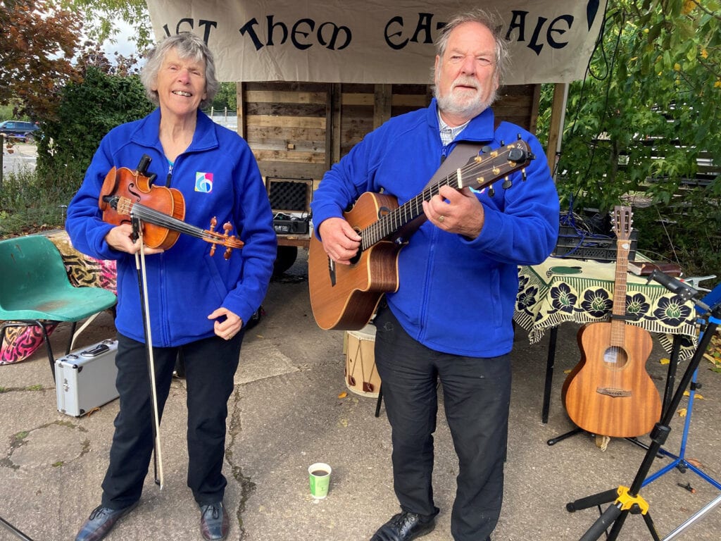 Musicians Peter with his guitar and Moira with her fiddle, both in Music in Hospitals & Care fleeces, standing in front of a sign reading 'Let them eat kale' busking.
