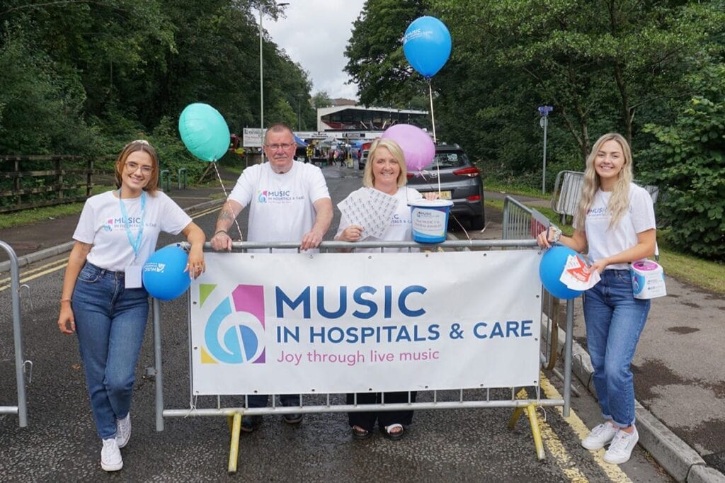Three women and one man stand around a Music in Hospitals & Care banner with balloons attached to it holding donation buckets.