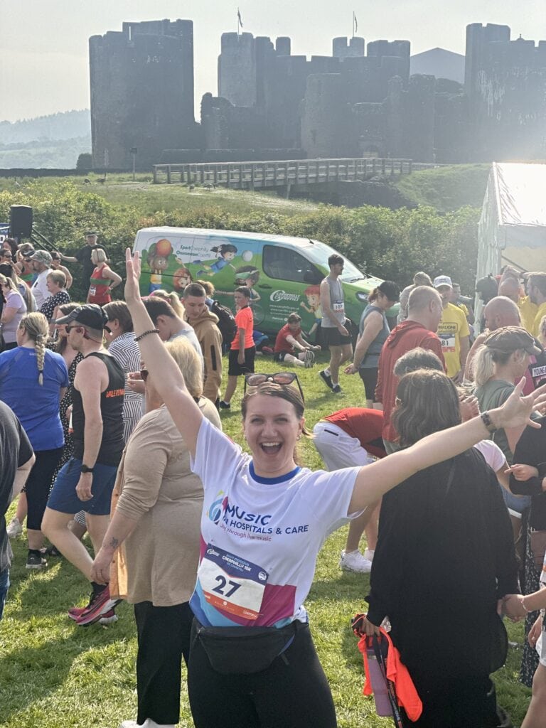A woman in a Music in Hospitals & Care running top with her arms in the air in front of Caerphilly castle.