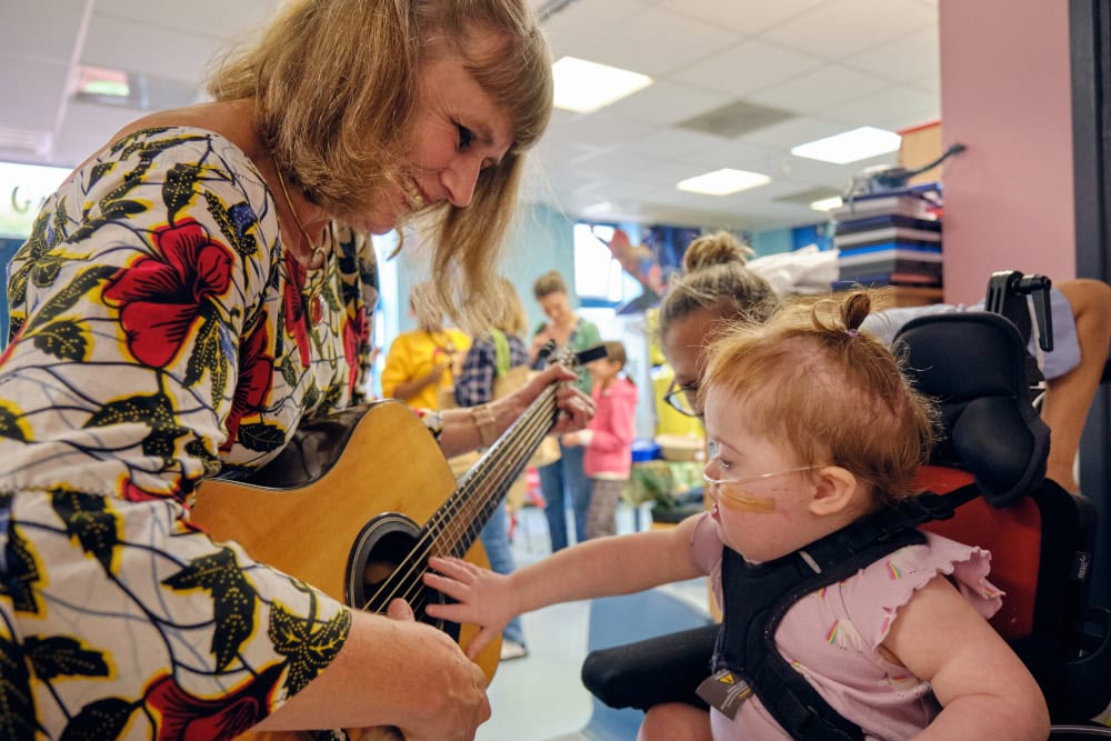 A woman holding a guitar smiling as a child in a special wheelchair with tube under her nose reaches out and strums the strings.