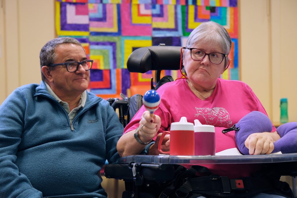 A lady in a wheelchair shakes a maraca. Her husband sits next to her smiling.