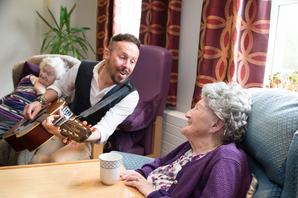 Musician playing guitar and singing to care home residents during a Music in Hospitals & Care session, with one woman smiling and engaging.