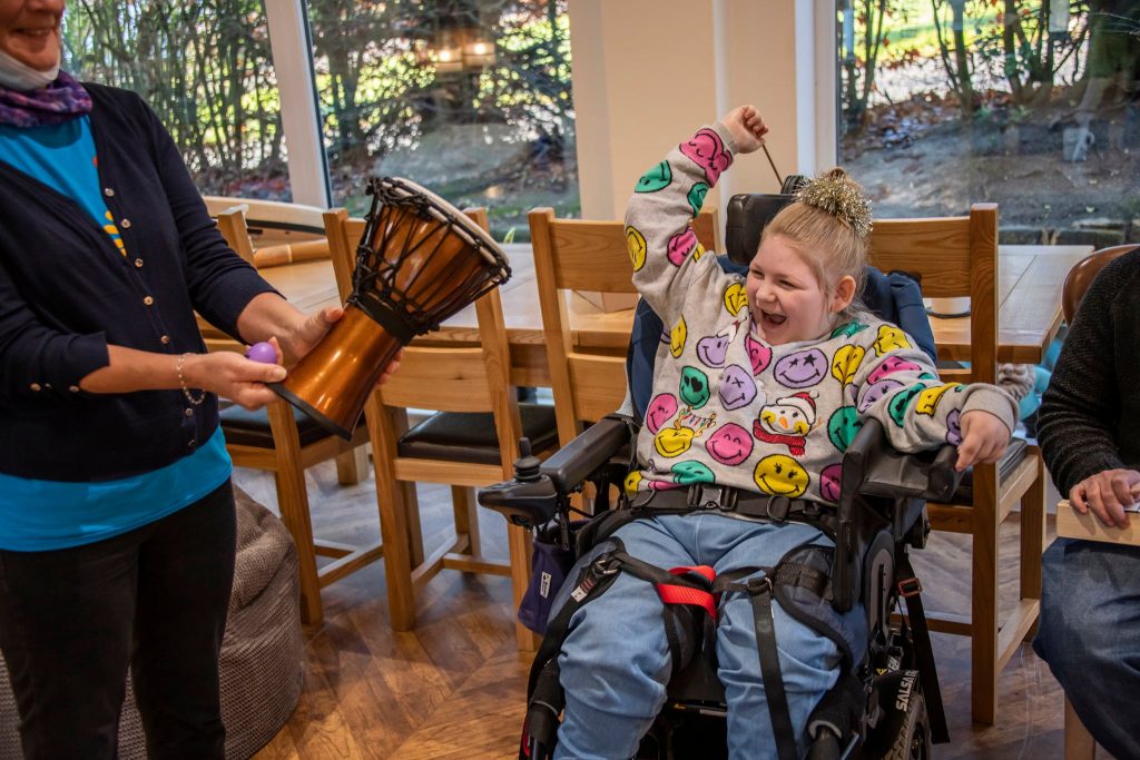 Child in a wheelchair smiling and raising their arm in excitement during an interactive drumming session with Music in Hospitals & Care.