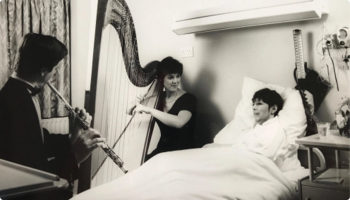 Historic photo of a harpist and flautist performing at a patient’s bedside in hospital as part of a Music in Hospitals & Care session.