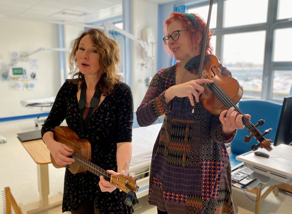Two musicians performing in a hospital ward, one playing ukulele and the other violin, during a Music in Hospitals & Care session.