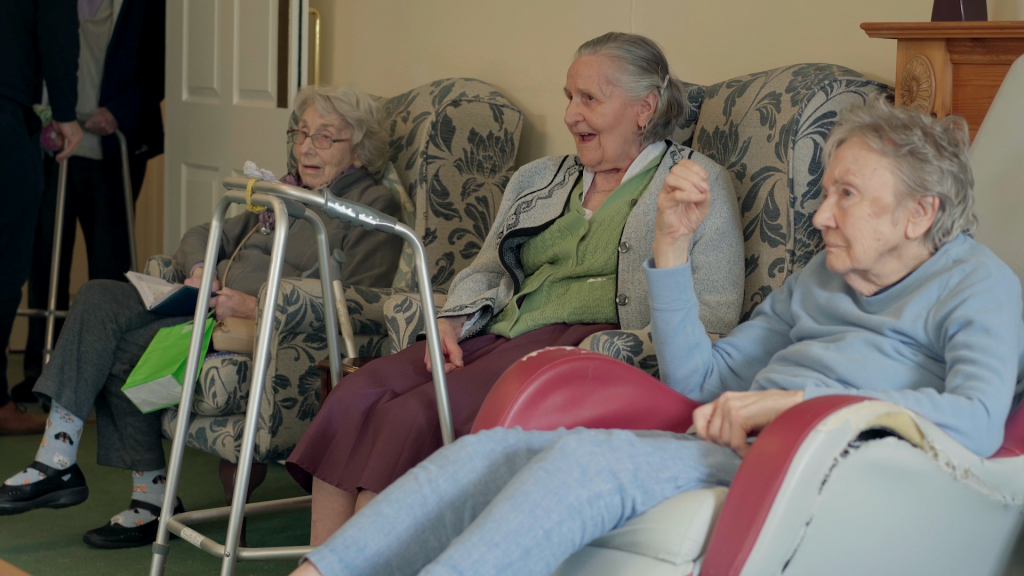 Elderly audience seated in a care-home lounge enjoying a live music performance by Music in Hospitals & Care