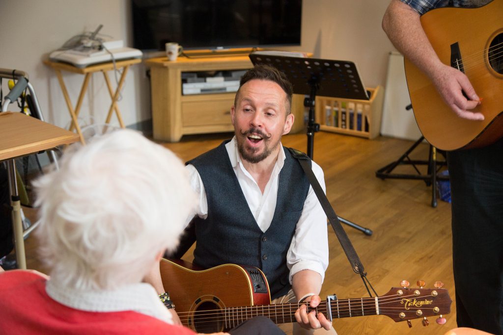 Musician playing guitar and singing to a care home resident during a Music in Hospitals & Care session.
