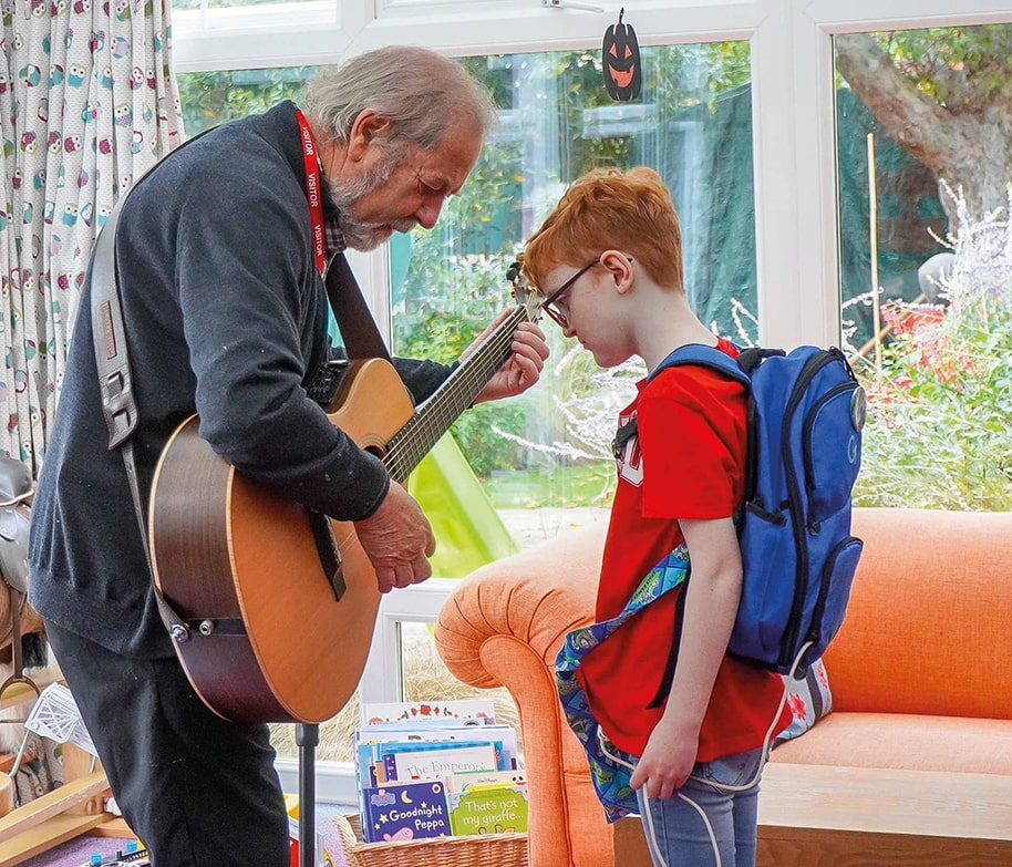 Musician playing guitar for a young boy with a backpack in a bright room, creating a warm and engaging moment.
