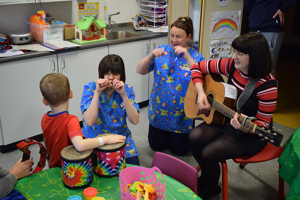 Hospital play specialists and a musician with guitar leading a joyful music session for a young child during a Music in Hospitals & Care visit.