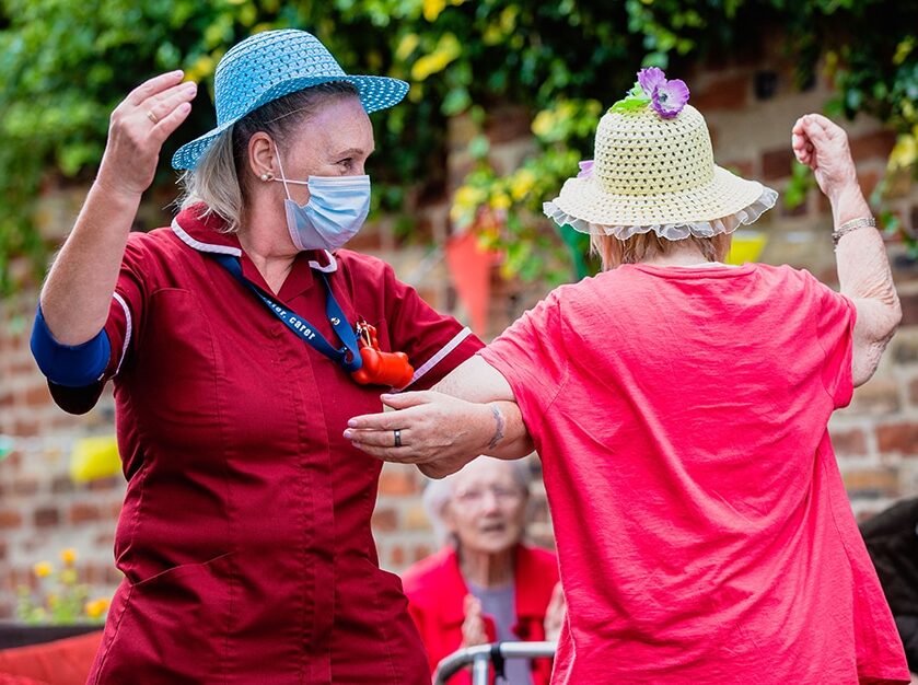 Carer and resident dancing together outdoors during a joyful care-home event, both wearing colourful hats.