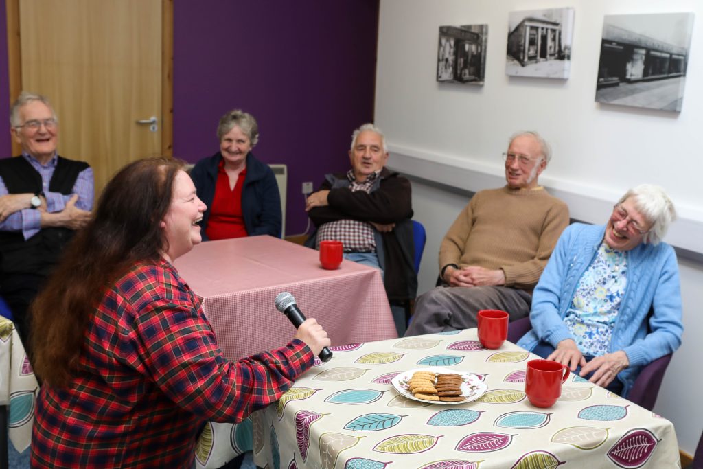 Performer singing with a microphone as older adults laugh and enjoy a Music in Hospitals & Care session over tea and biscuits.