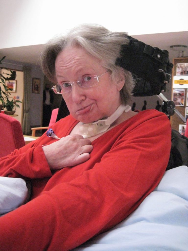 Woman in a red top seated in a care setting, looking toward the camera with a gentle expression.