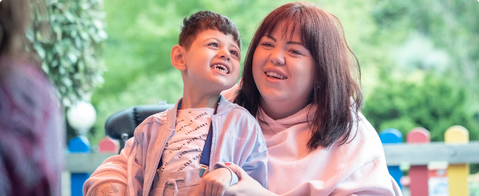 Parent smiling with their young child in a wheelchair during an outdoor Music in Hospitals & Care event.