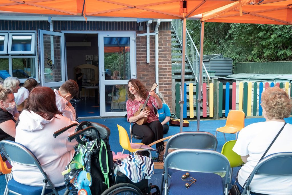 Musician playing ukulele for children and families during an outdoor Music in Hospitals & Care session under a bright orange canopy.