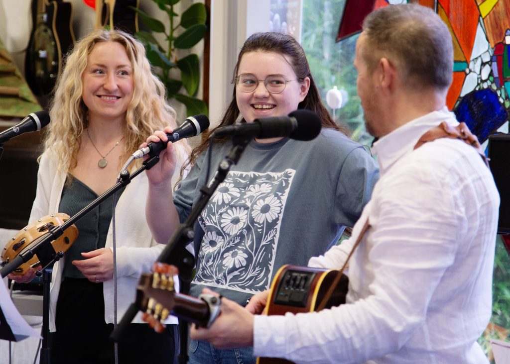Three musicians smiling and performing together with microphones and guitar during a Music in Hospitals & Care session.