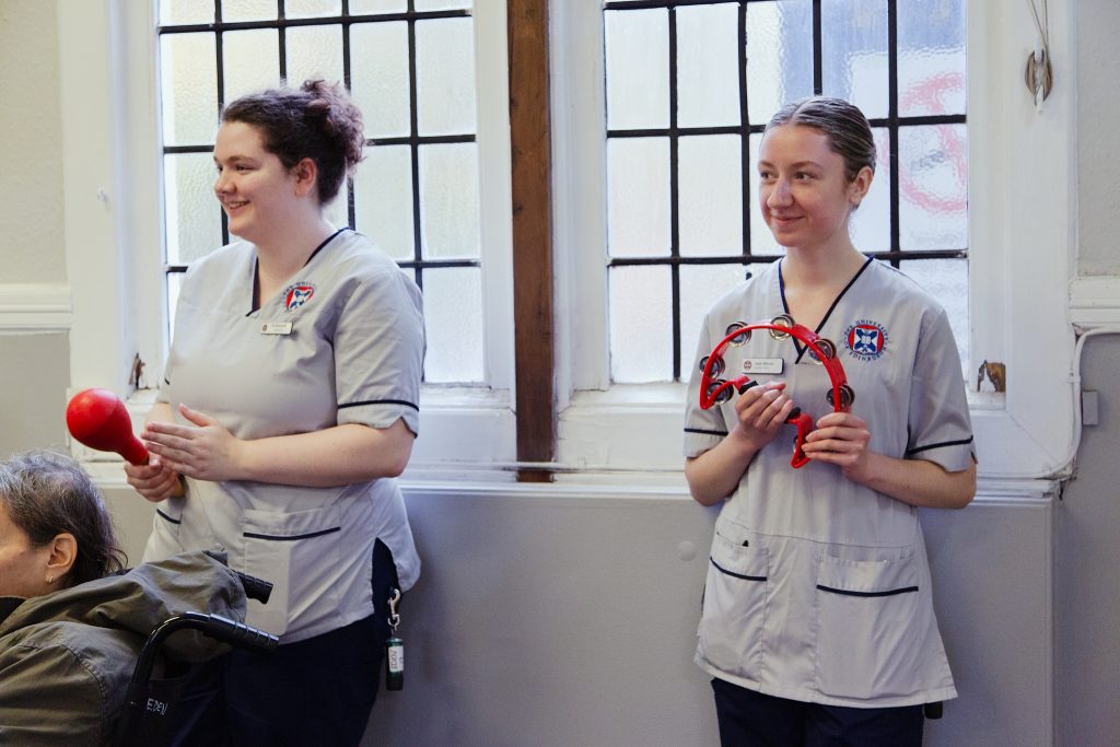 Two healthcare staff smiling and playing percussion instruments during a Music in Hospitals & Care session.