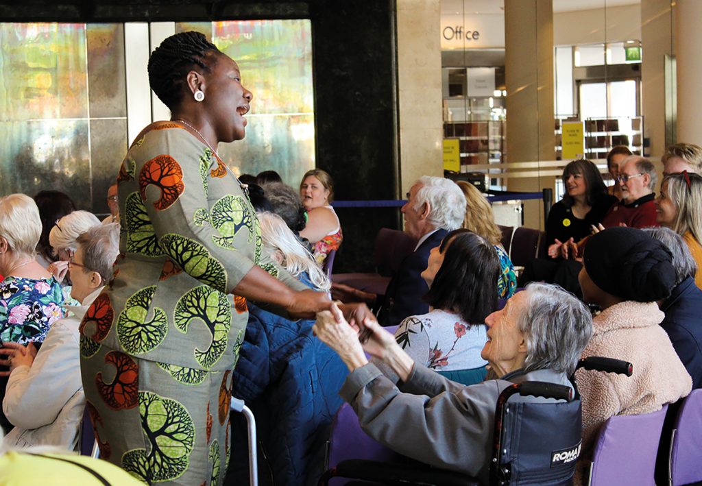 Performer singing and holding hands with an audience member in a wheelchair during a Music in Hospitals & Care live event.