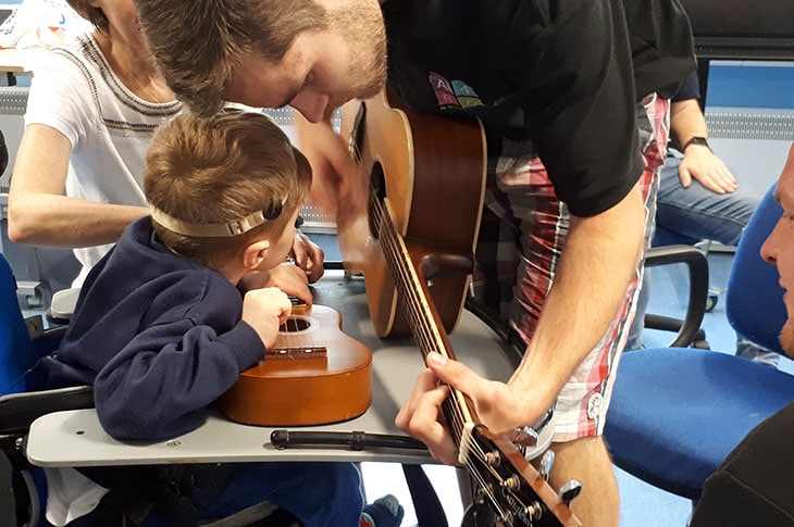 Musician helping a young child with hearing aids strum a guitar during an interactive Music in Hospitals & Care session.