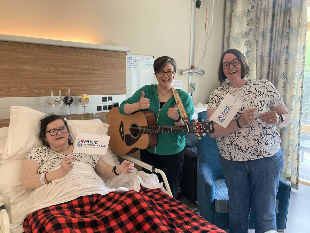 Musician with guitar performing at a patient’s bedside while two women hold Music in Hospitals & Care signs and smile for the camera.