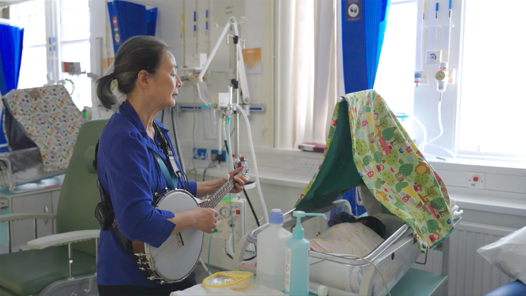 Musician playing banjo at a hospital bedside for a baby in intensive care during a Music in Hospitals & Care session.