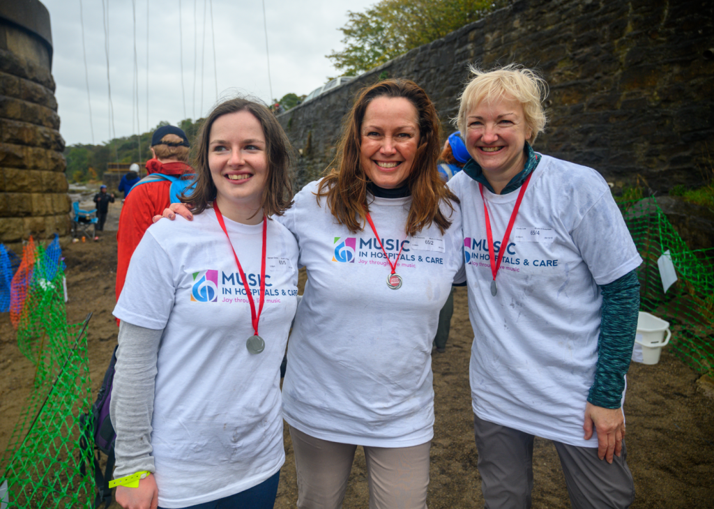Three smiling women wearing ‘Music in Hospitals & Care’ T-shirts and medals after a fundraising event.
