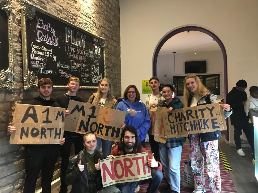 Group of smiling participants holding cardboard signs for a charity hitchhike event with messages such as ‘M1 North’ and ‘Charity Hitchhike.