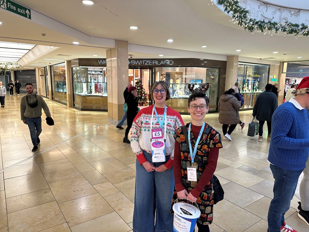 Two smiling fundraisers wearing festive outfits and ‘Music in Hospitals & Care’ lanyards inside a shopping centre decorated for Christmas.