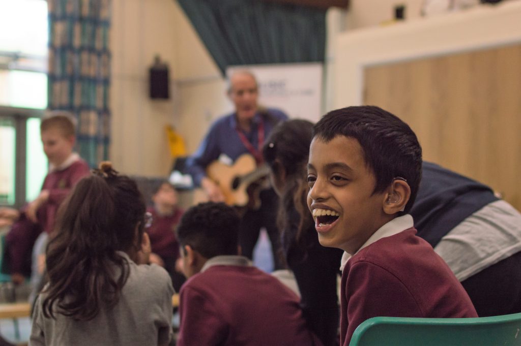 Smiling schoolboy enjoying a live music session led by a guitarist in a classroom with other children.