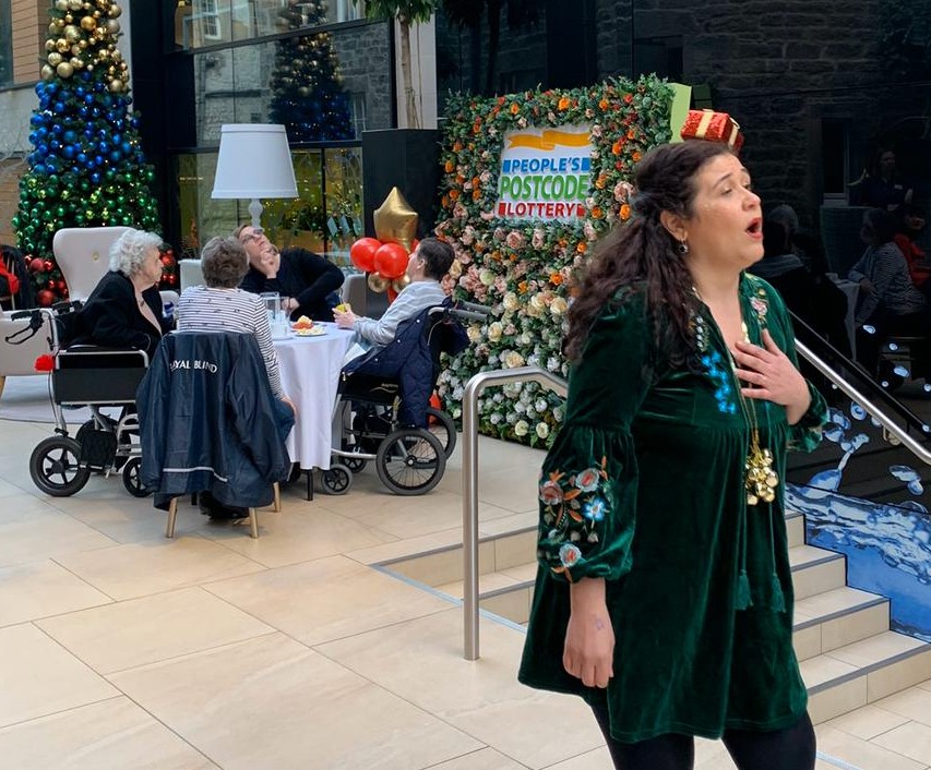 Performer singing at a festive event with audience members seated nearby and a People’s Postcode Lottery floral display in the background.