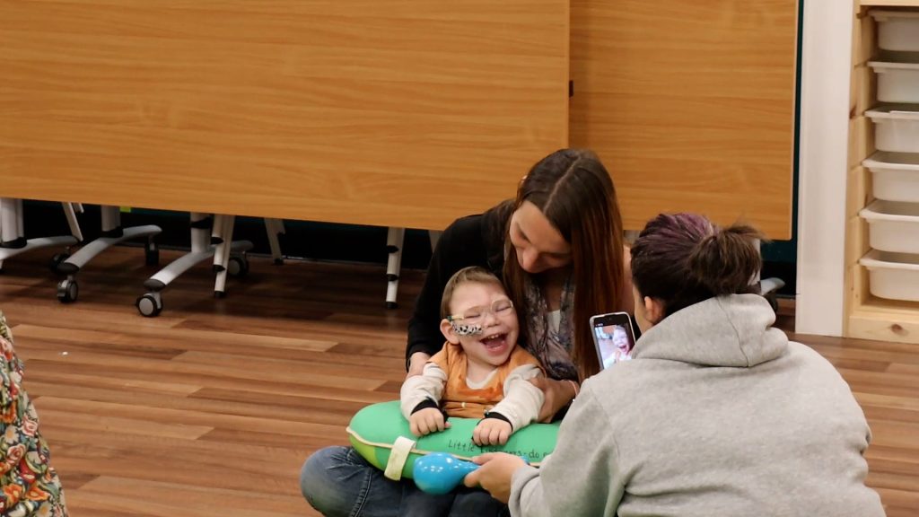 Young child with medical support laughing while engaging in a Music in Hospitals & Care session with two adults.
