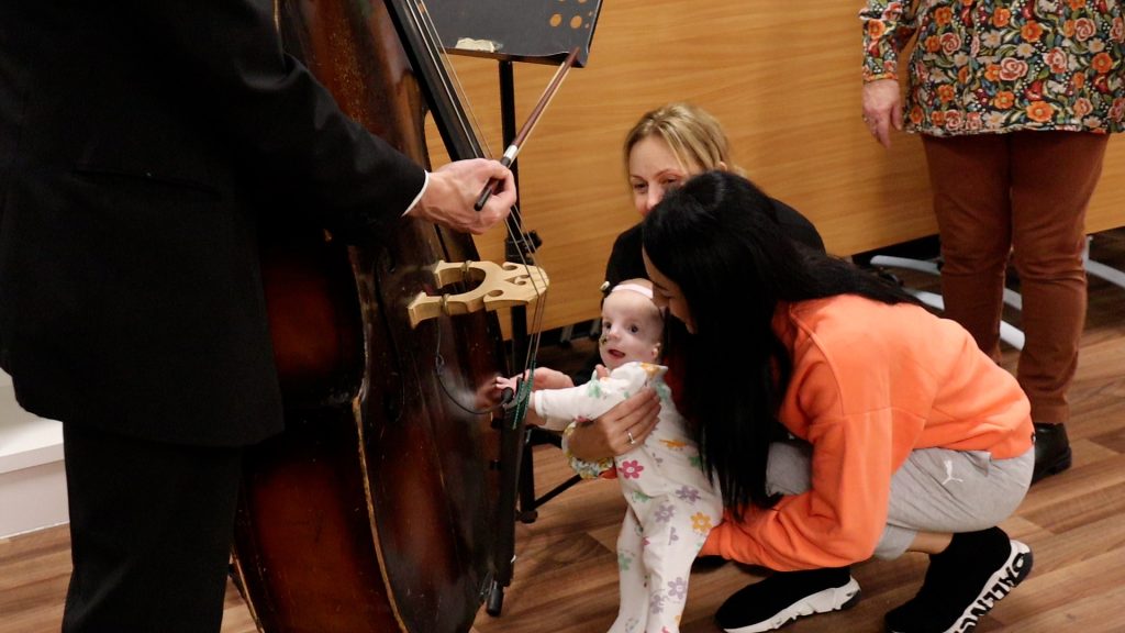Baby reaching out to touch a double bass during an interactive Music in Hospitals & Care session, supported by a parent