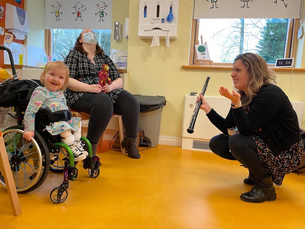 Musician playing clarinet for a smiling child in a wheelchair during a Music in Hospitals & Care session, with a parent watching.