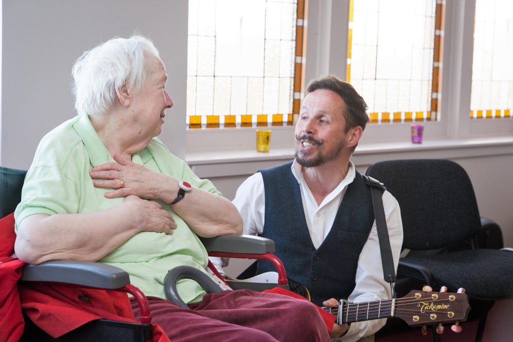 Musician playing guitar and singing to care home residents during a Music in Hospitals & Care session, with one woman smiling and engaging