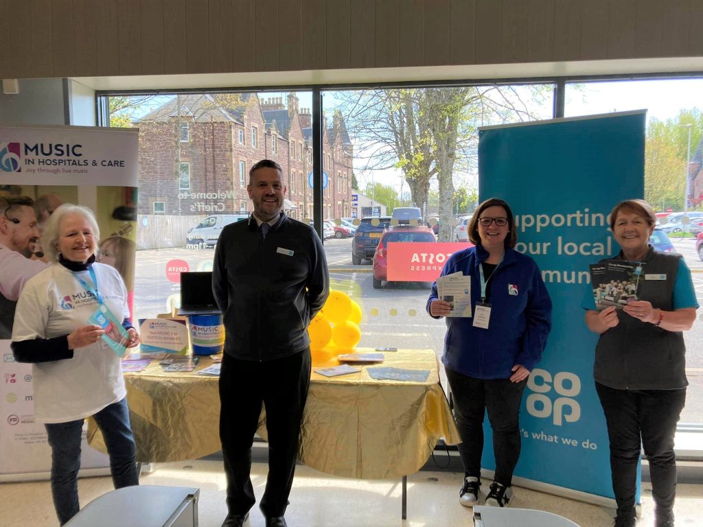 Representatives from Music in Hospitals & Care and Co-op standing together at a community fundraising event with banners and leaflets.