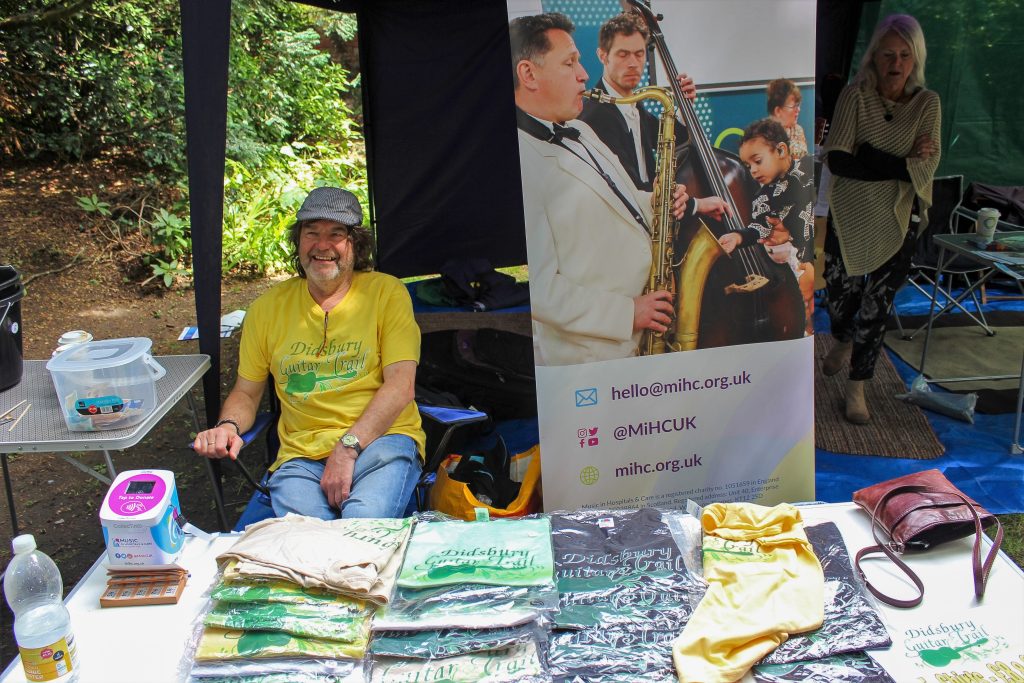 Person smiling at an outdoor fundraising stall with T-shirts on display beside a Music in Hospitals & Care banner.