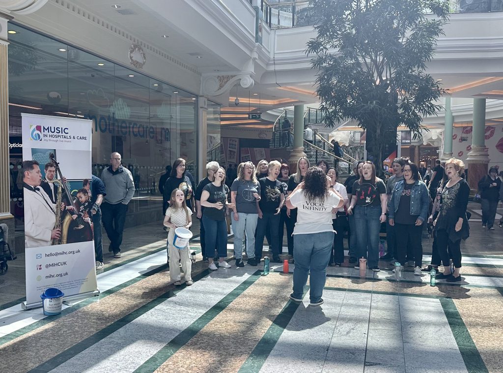 Choir performing in a shopping centre with a conductor leading them and a ‘Music in Hospitals & Care’ banner displayed beside donation buckets.