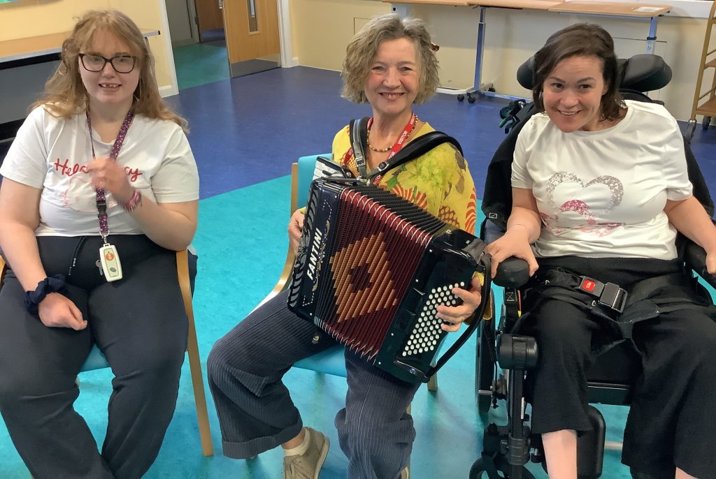 Accordionist performing alongside two smiling women during an inclusive Music in Hospitals & Care session.