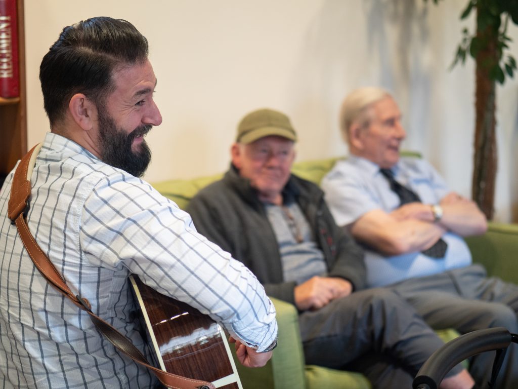 Musician smiling with guitar while engaging two older men during a Music in Hospitals & Care session.