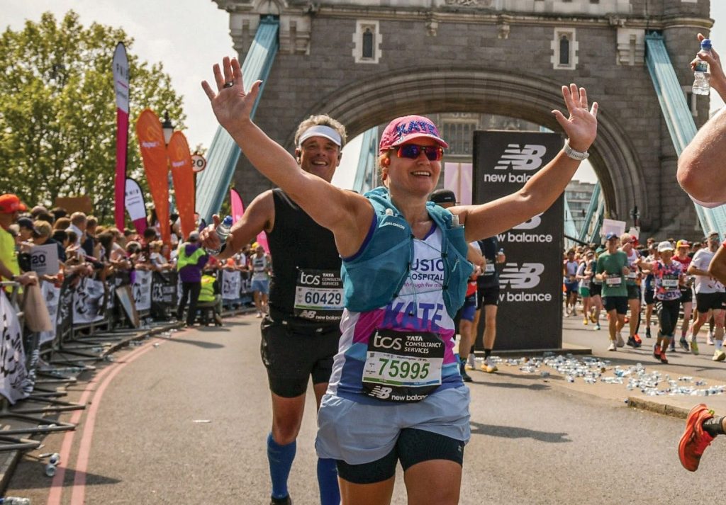 Runner wearing a Music in Hospitals & Care vest smiles and raises her arms while crossing Tower Bridge during a marathon.