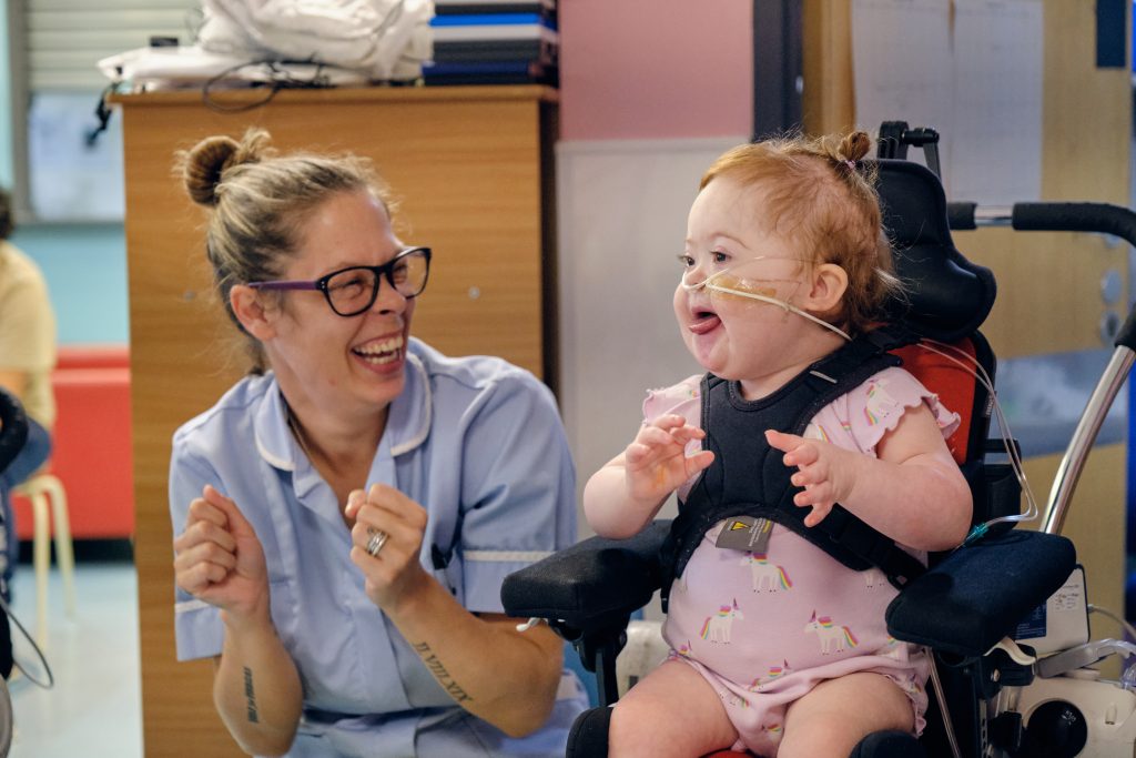 Nurse smiling and clapping with a young child in a wheelchair enjoying live music during a Music in Hospitals & Care session.