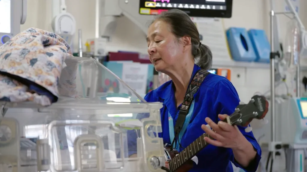 Musician playing banjo beside an incubator for a baby in neonatal care during a Music in Hospitals & Care session.