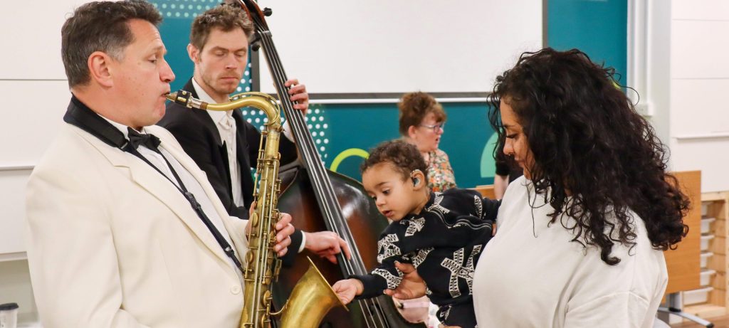 Musicians performing jazz while a parent helps a young child with hearing aids explore the sound of a saxophone during a Music in Hospitals & Care session.