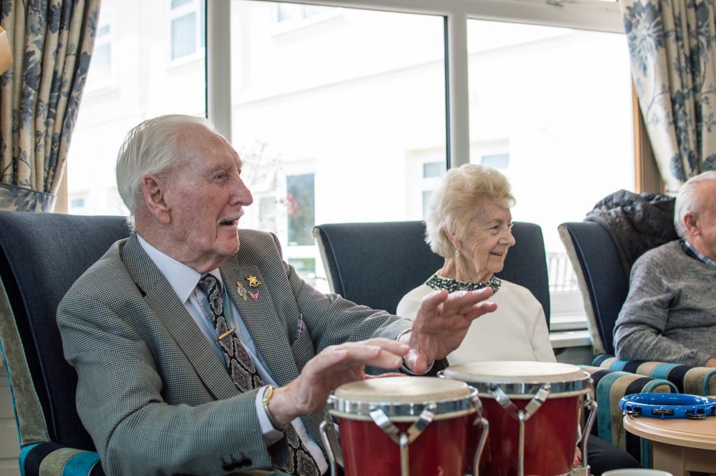 Elderly man and woman smiling and playing drums together during a live music session in a care home.