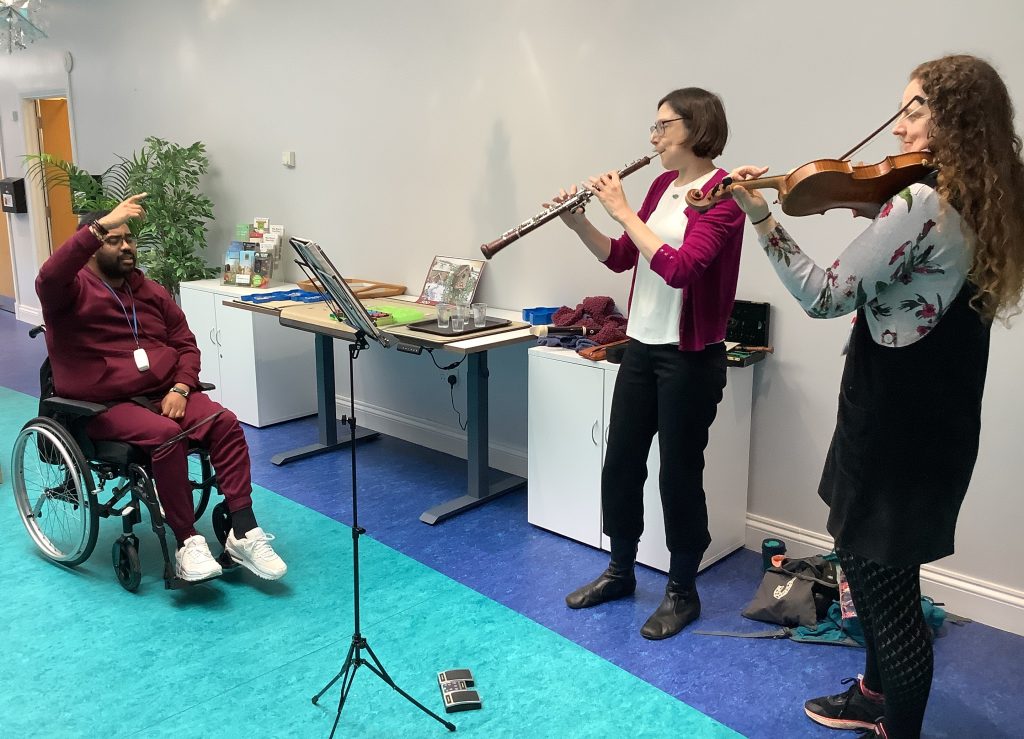 “Two musicians performing on flute and violin for a man in a wheelchair, who raises his hand joyfully in response to the live music.