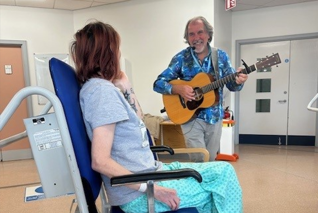 Musician playing guitar and singing to a hospital patient during a Music in Hospitals & Care bedside performance.