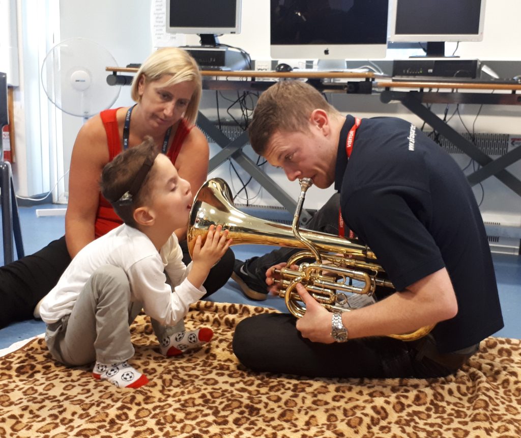 Musician helping a young child explore the sound of a brass instrument during a Music in Hospitals & Care interactive session, with carer watching nearby.